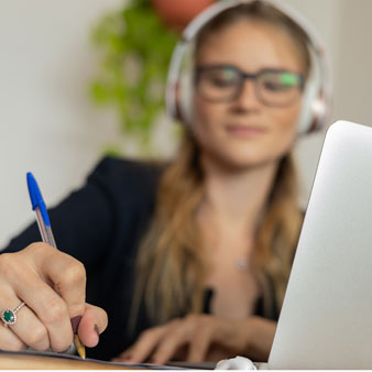 Mujer con auriculares y gafas escribe con un bolígrafo frente a un portátil